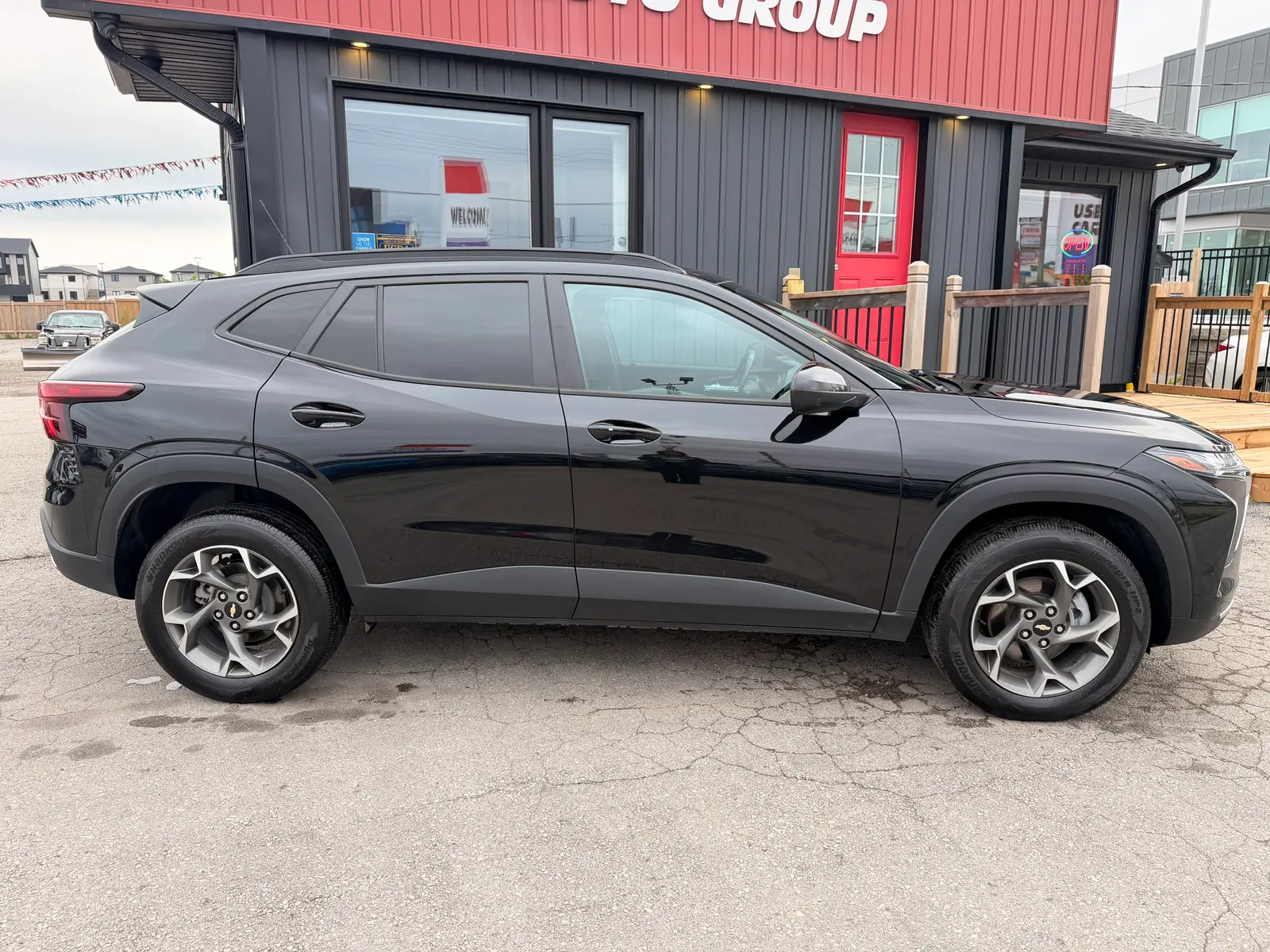 2024 Chevrolet Trax 1LT Black driver side profile view with multi-spoke alloy wheels and dealership storefront background.