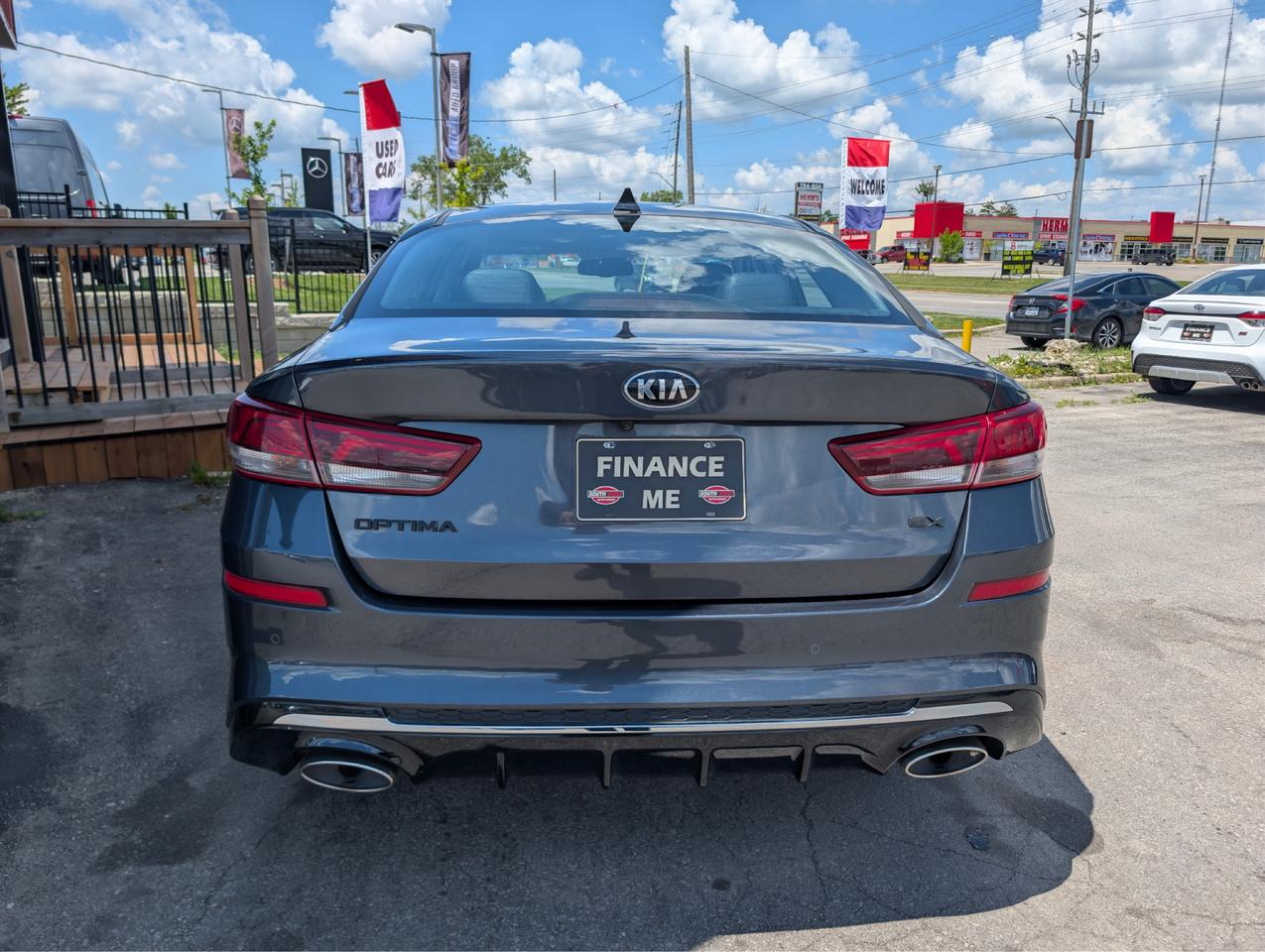 2020 Kia Optima Sedan Gray rear exterior view showing dual exhaust outlets, red taillights, and dealership lot setting.